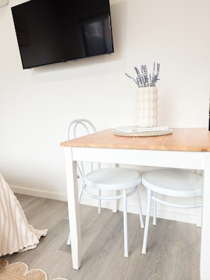 A dining area with a wooden table, white chairs, a television mounted on the wall, and a vase with plants on the table.