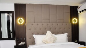 A hotel room with a tufted headboard, a bed with white bedding and an orange throw, a nightstand with a phone, and a wall-mounted air conditioner. - Deedee Luxury Apartments (Osogbo)