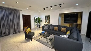 A modern living room with a sofa, coffee table, and a chair. There is a large window with blinds, a door, and a plant. - Deedee Luxury Apartments (Osogbo)