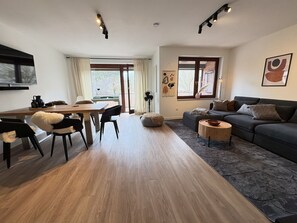 A modern living room with a wooden floor, a dark grey sofa, a round coffee table, and a dining area with a wooden table and chairs.