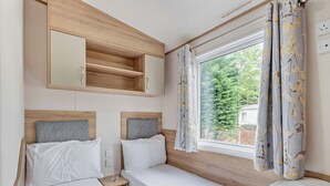 A double bed room with a wooden headboard, a bedside table, a mirror, and a window with curtains. - Gold Caravan with parking & decking P (Walton-on-the-Naze)