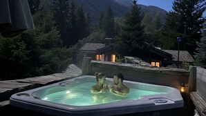 Ein Whirlpool mit zwei Personen, umgeben von einer Berglandschaft und Holzhäusern.