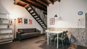 Een woonkamer met een bank, een eettafel met stoelen, een trap en een muur met een patroon betegelde vloer en een klok.