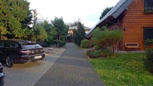 Ein gepflasterter Weg führt zu einem Holzhaus mit Satteldach, umgeben von Grünflächen und einem parkenden Auto.
