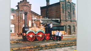 Eine Dampflokomotive mit roten Rädern, ein Backsteingebäude und eine Gruppe Personen davor.