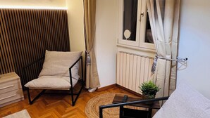 Un dormitorio moderno con cama, una silla, una mesita con planta, una lámpara y un ventanal con cortinas.