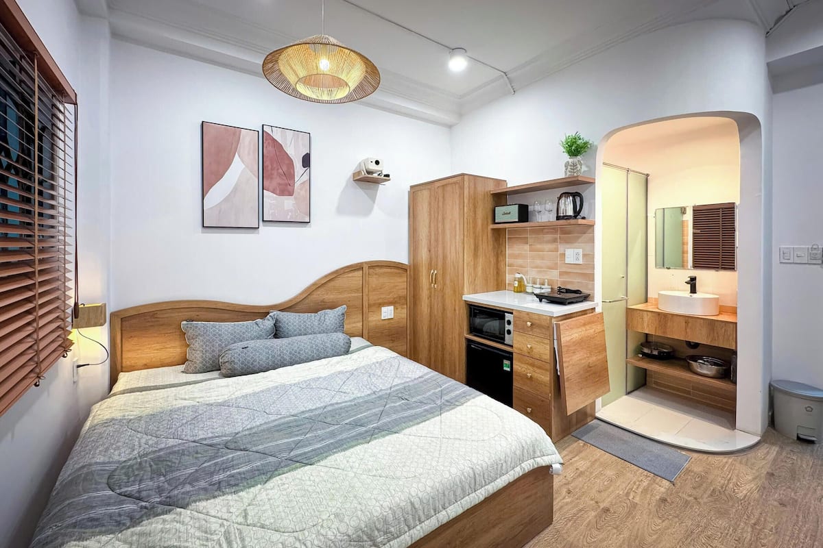 Un dormitorio con cabecera de madera, cama con ropa de cama estampada, mesita de noche, armario, zona de cocina integrada con fregadero y encimera, y un espejo.