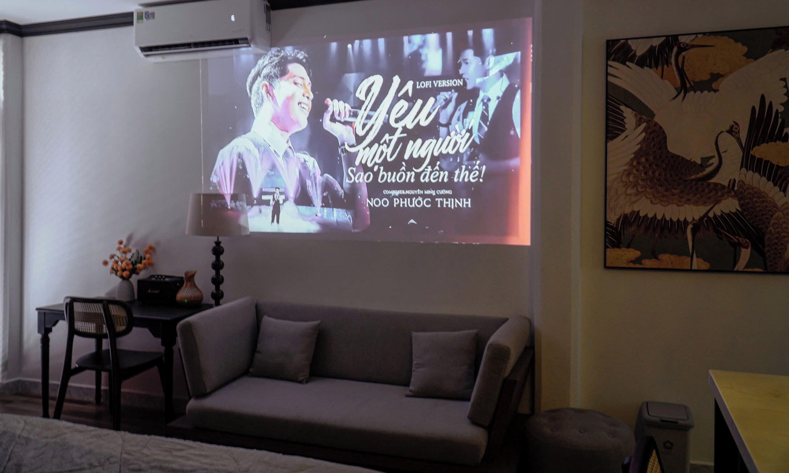 A living room with a sofa, a small table with a lamp, and a wall-mounted TV displaying a song.