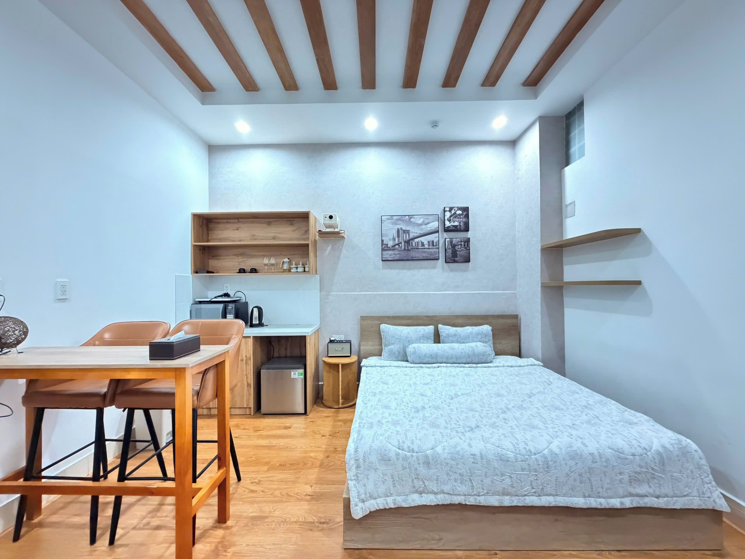 A compact hotel room with a bed, a desk with a chair, a small kitchenette, and a shelf with pictures.