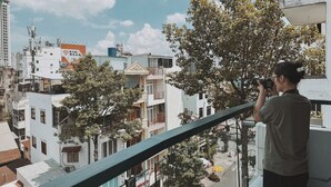 Une personne prend une photo depuis un balcon donnant sur un paysage urbain, avec des bâtiments et des arbres.