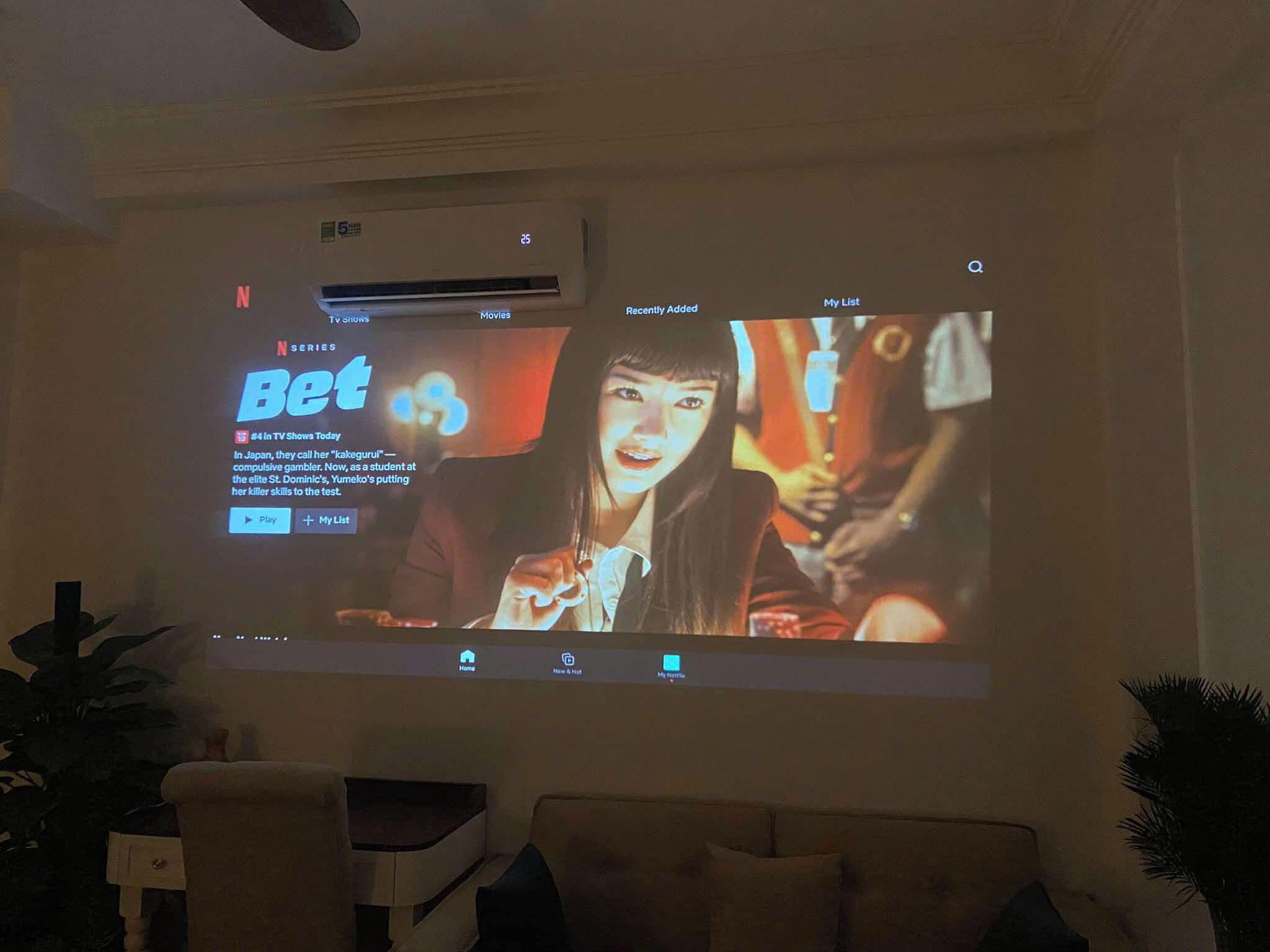 Una habitación con una pantalla grande mostrando una película o programa de televisión, un sofá y una planta.