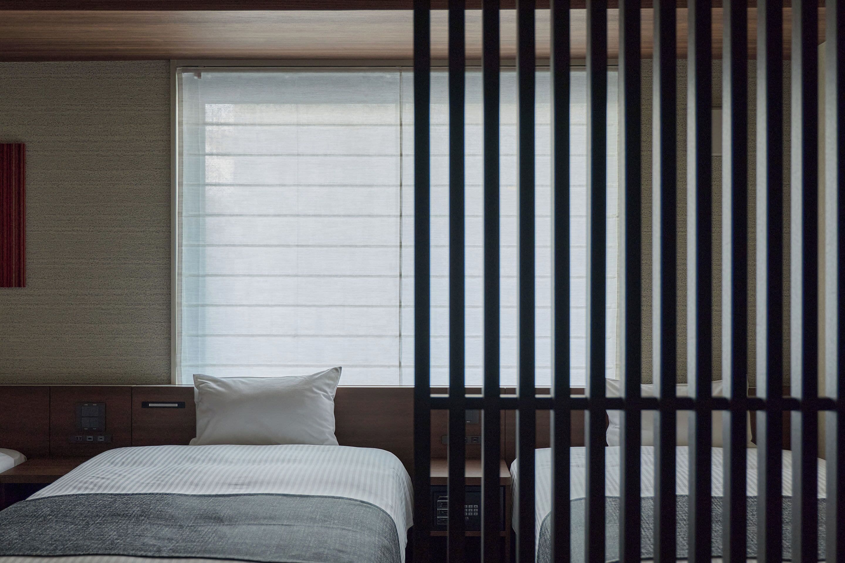 A hotel room with a bed, a bedside table, a window with blinds, and a view of the outside.