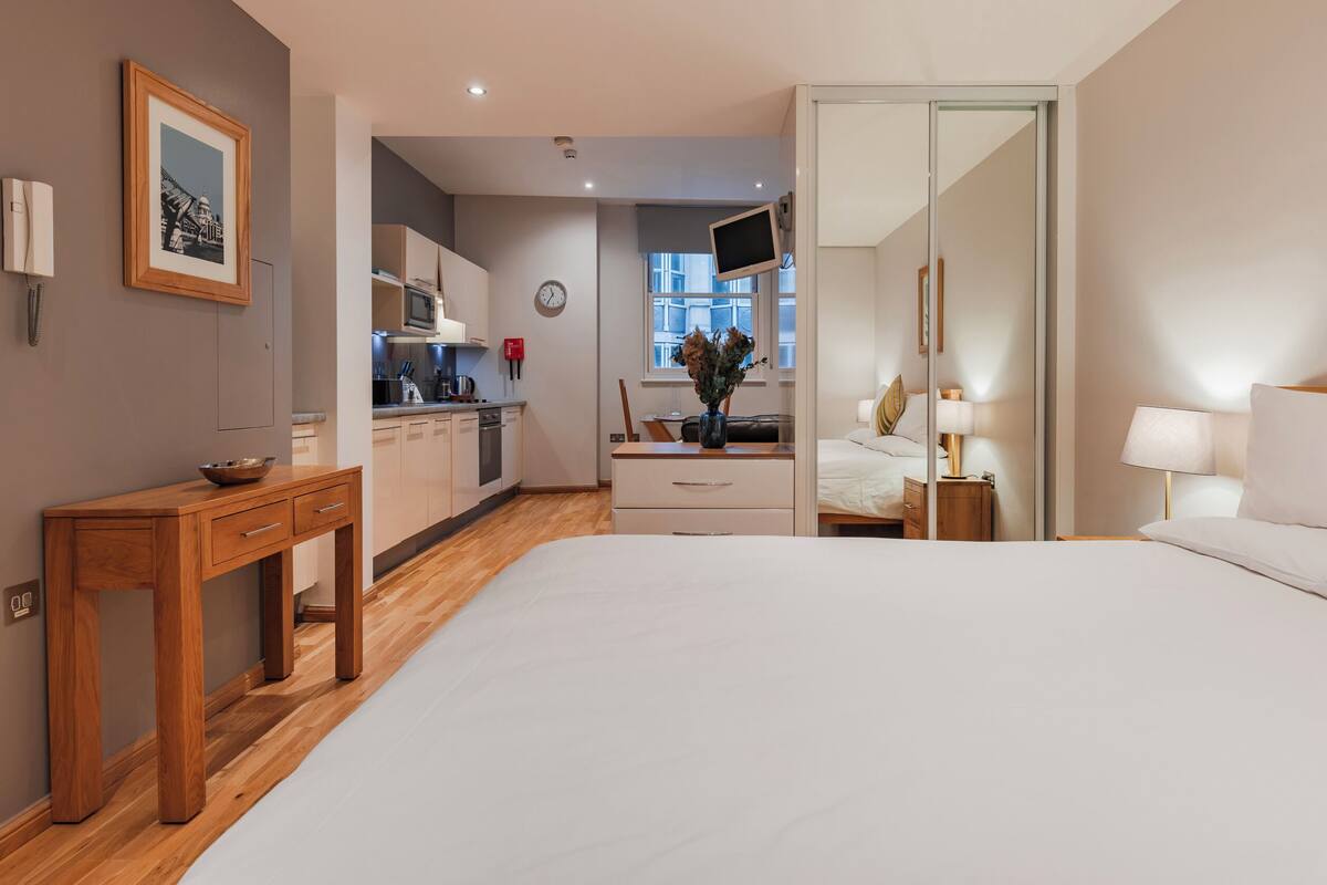 A hotel room with a bed, bedside table, a mirror, a TV, a small kitchen area, and a view of the city.