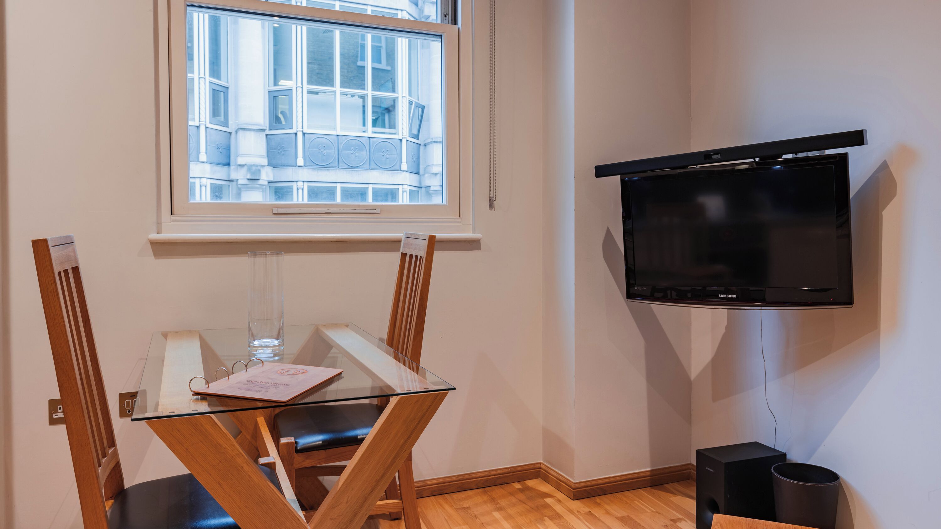 A room with a dining table, chairs, a television mounted on the wall, and a window with a view of buildings.