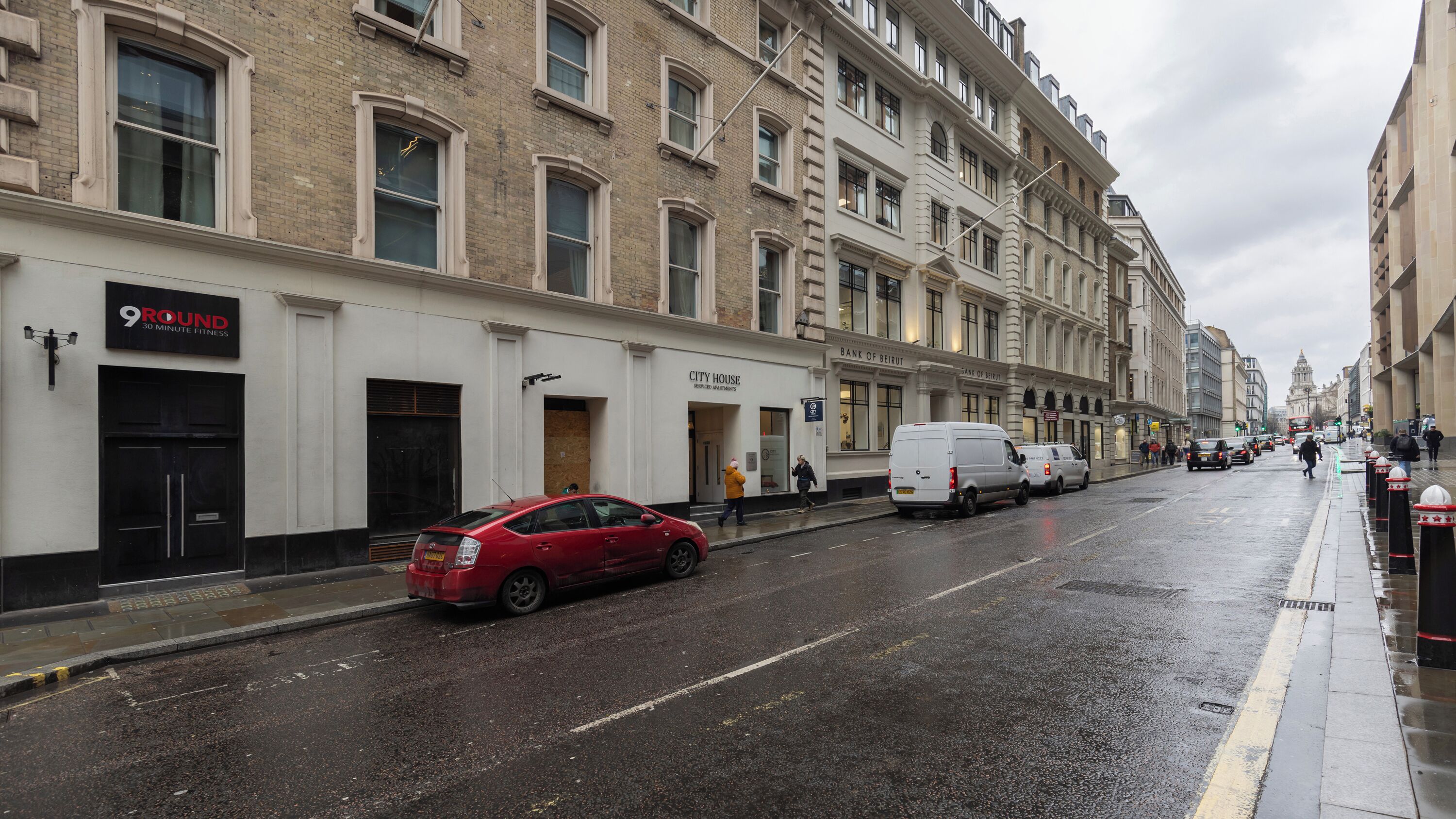 Une rue où est garée une voiture rouge sur le bord, un fourgon blanc, et un immeuble avec une enseigne indiquant « City House » et « Bank of Beirut ».