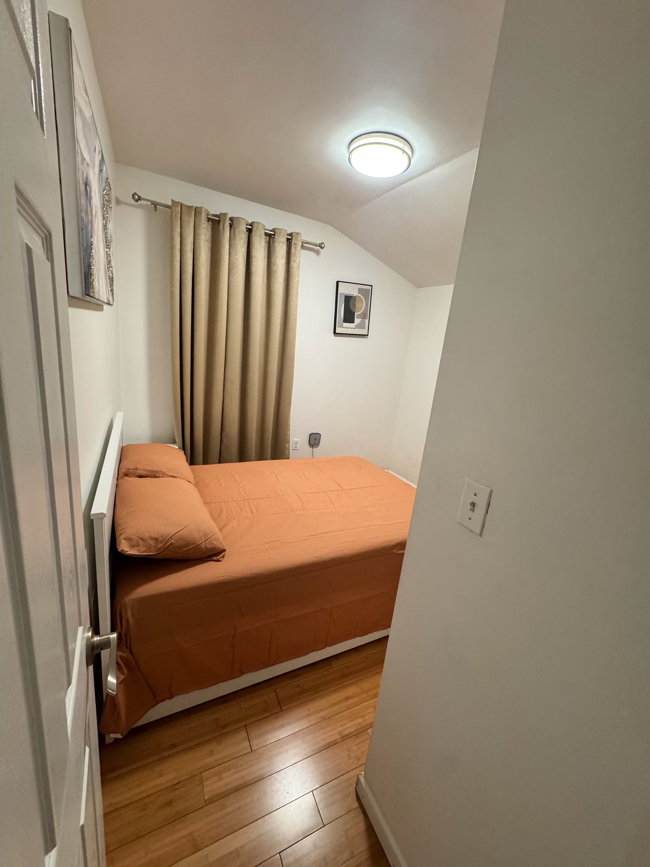 A bedroom with a bed, curtains, a ceiling light, a wall-mounted picture, and a wooden floor.