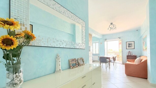 Ein Zimmer mit hellblauer Wand, einem weißen Schminktisch, einem Spiegel, einer Vase mit Sonnenblumen, einem weißen Leuchtturm und einem Essbereich mit Tisch und Stühlen.