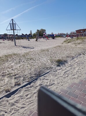 Uma praia arenosa com equipamentos de playground, incluindo escorregadores e balanços, e um edifício ao fundo.