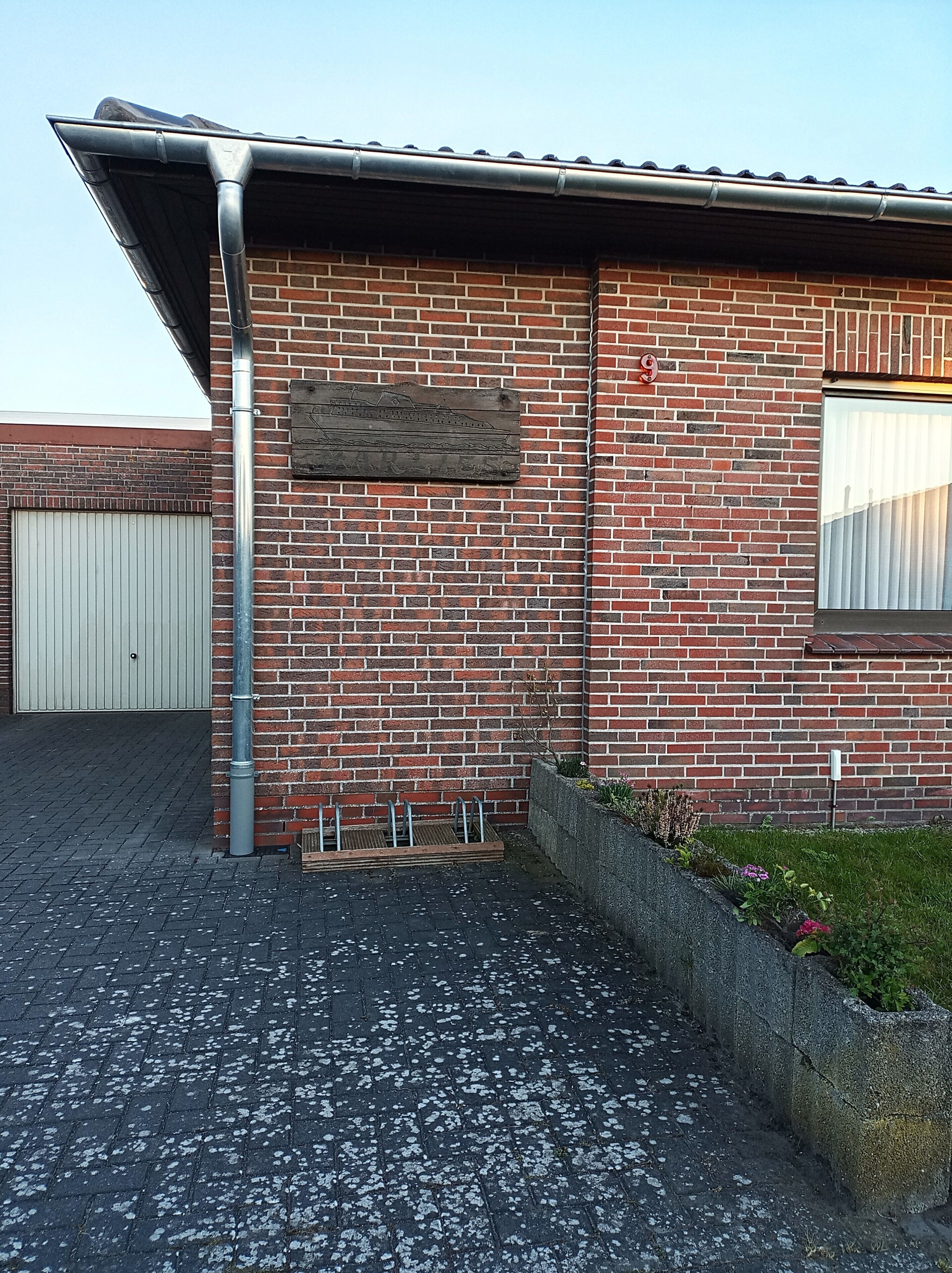 Un edificio de ladrillo con canalón metálico, puerta de garaje y una jardinera con plantas.