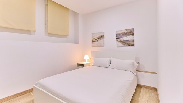 A bedroom with a bed, a nightstand, a wall-mounted lamp, and two abstract paintings.