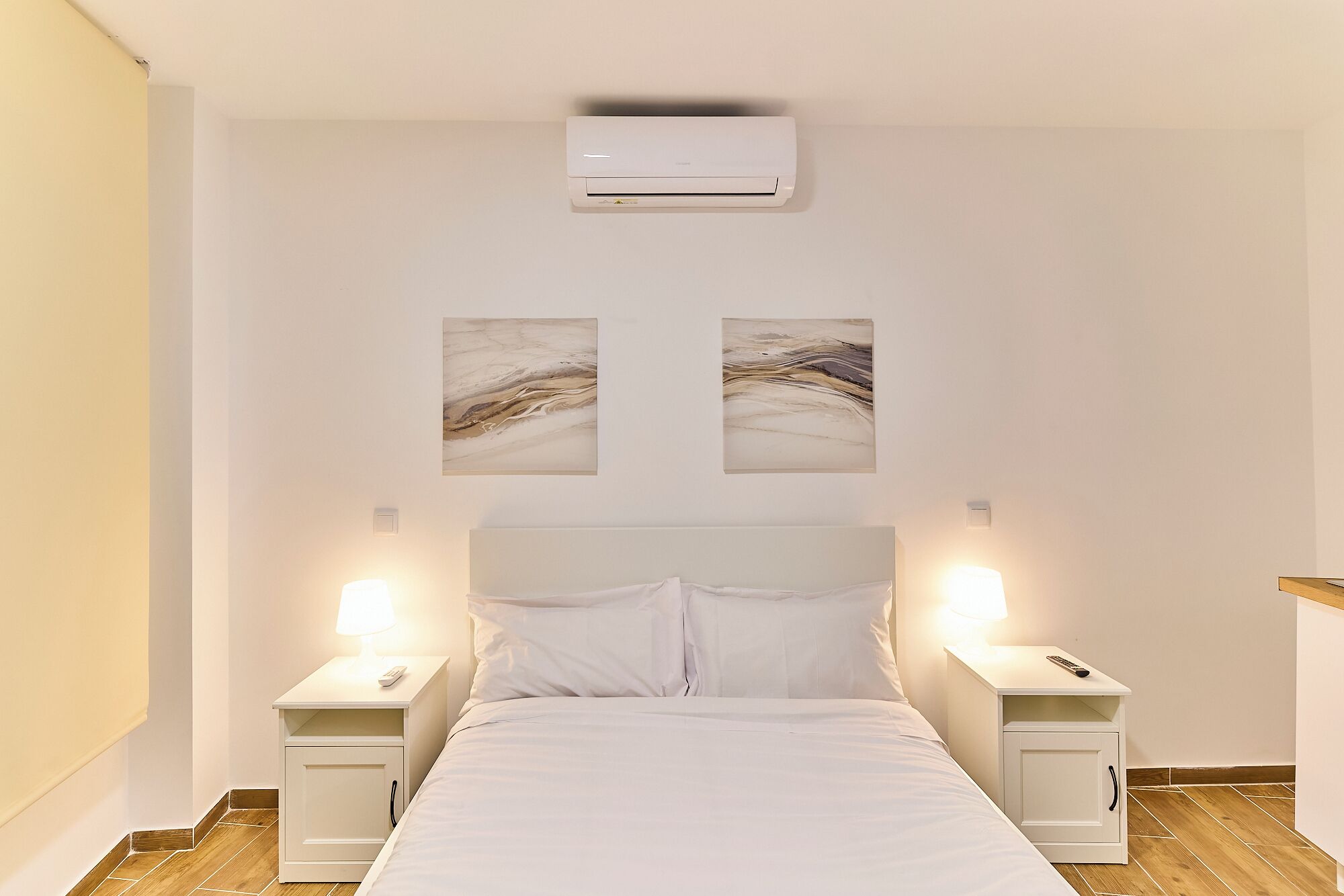 Een slaapkamer met een bed, twee nachtkastjes met lampen, een airconditioner en twee abstracte kunstwerken aan de muur.