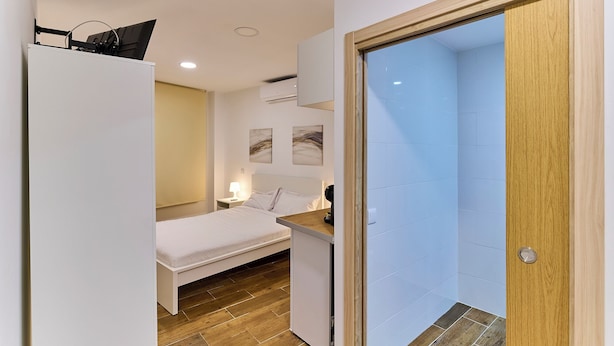Quarto pequeno com cama, mesa de cabeceira e porta de madeira que dá para um banheiro.