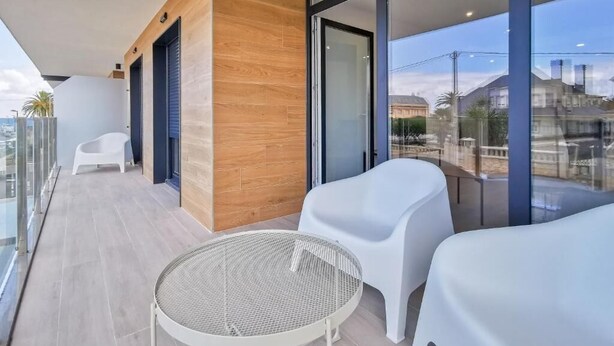 A balcony with a round table and two white chairs, a wooden accent wall, and a glass door leading to a view of buildings. - Apartamentos Costa de Foz by gaiarooms  (Foz)