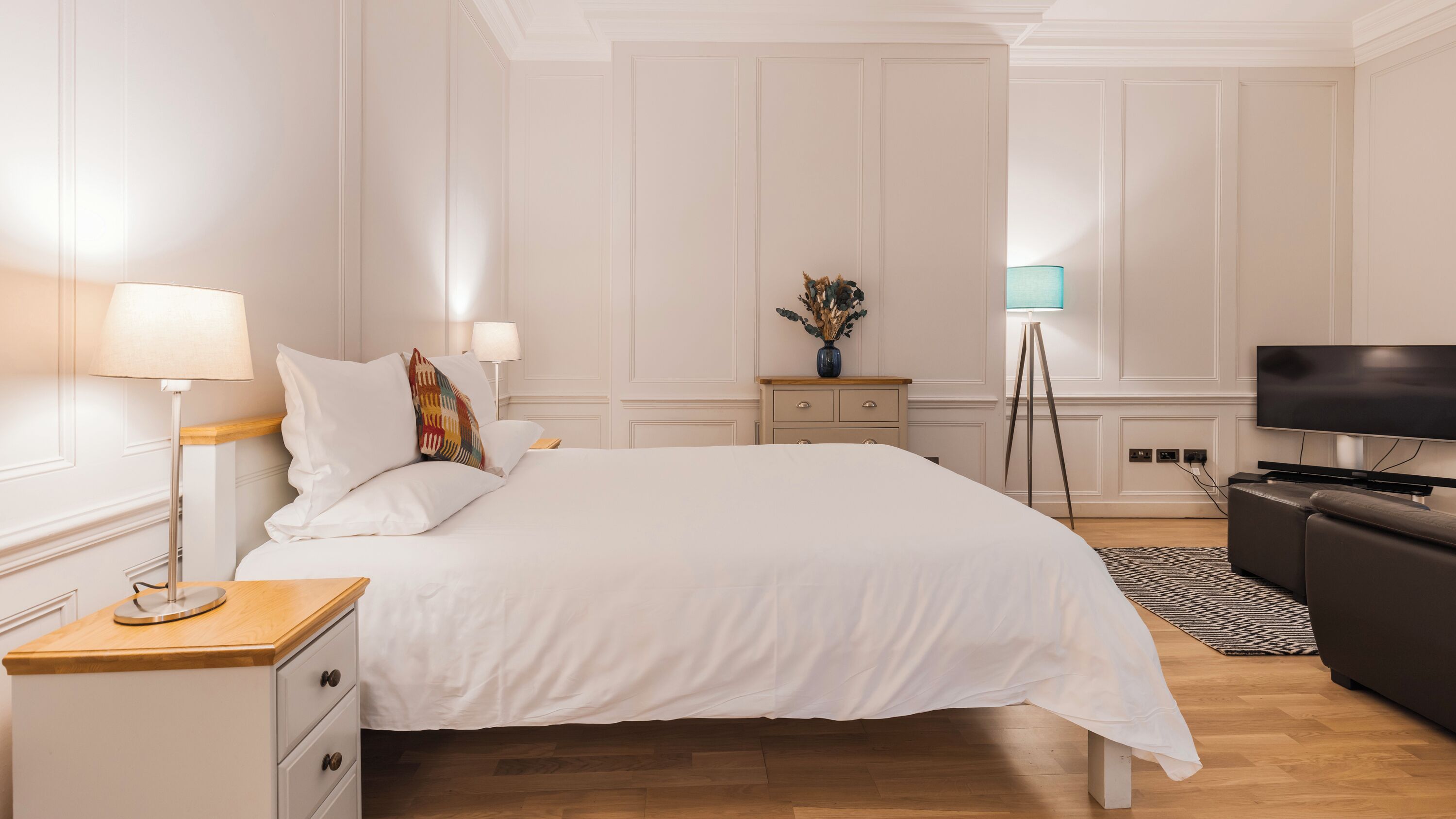 Hotellihuone, jossa on sänky, yöpöydät, työpöytä, televisio ja lattiavalaisin.