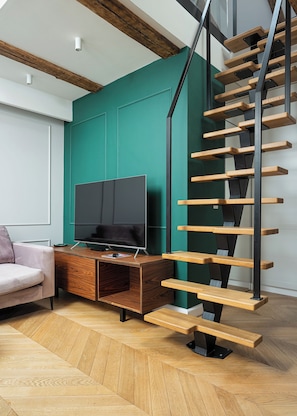 A modern living room with a wooden staircase, a flat-screen TV on a wooden console, and a teal accent wall. - RentPlanet - Piotrkowska (Łódź)