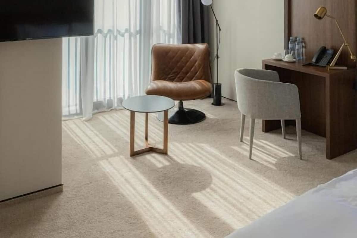 Habitación de hotel con cama, escritorio con lámpara, silla, televisor y una ventana grande con cortinas.