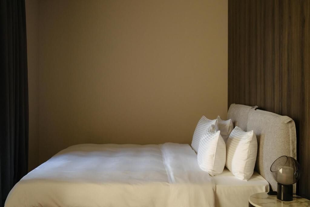 A neatly made bed with white bedding and pillows, a bedside table with a lamp, and a dark curtain on the left.