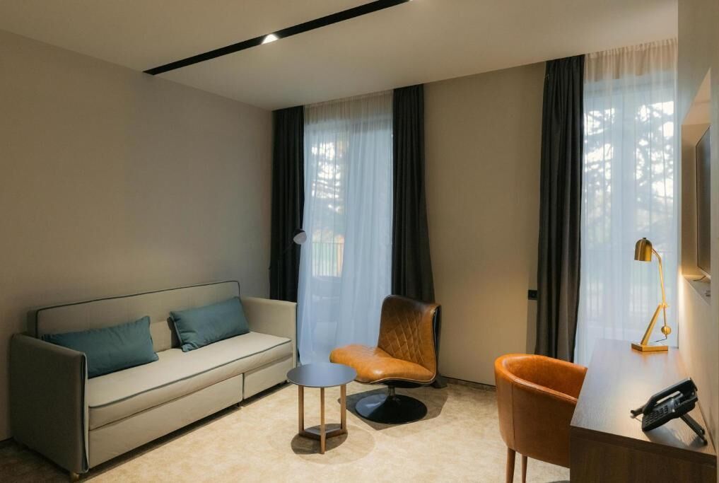 A modern hotel room with a sofa, a chair, a desk with a phone, and a lamp.