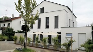 Een modern wit gebouw met veel ramen, een klein patio met een parasol, en een boom op de voorgrond.