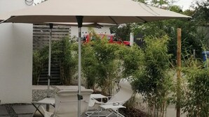 Een terras met een tafel en stoelen onder een grote parasol, omringd door groen en een houten schutting.