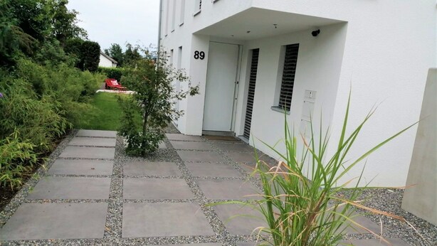 Een pad dat leidt naar een wit gebouw met nummer 89, omgeven door groen en een kleine plant in een pot.