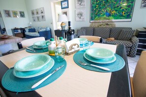 Tavola apparecchiata con piatti azzurri, posateria e una bottiglia, circondata da cuscini decorati e un quadro floreale incorniciato.
