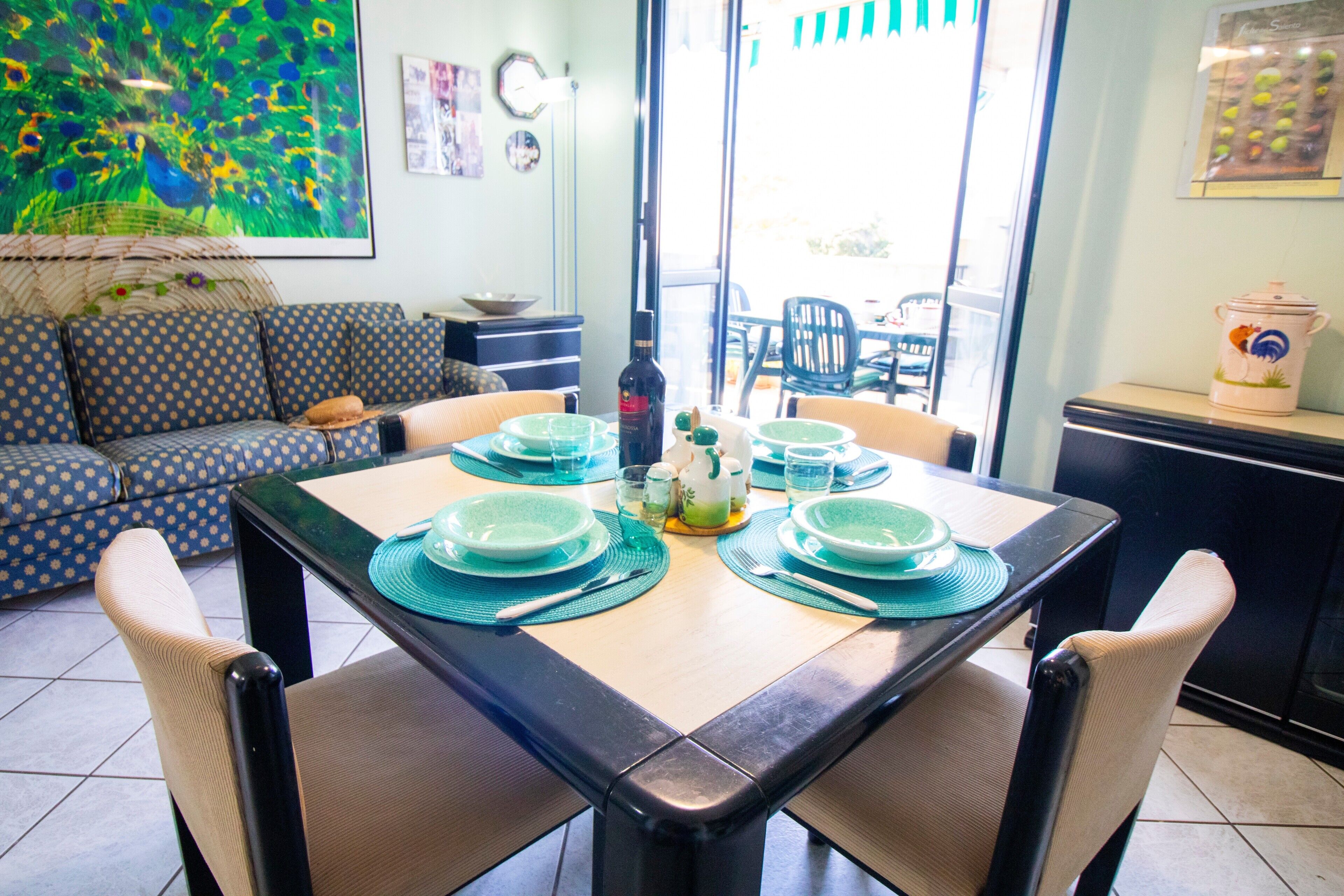 A dining area with a table set for four, chairs, a sofa, and a kitchen area in the background.