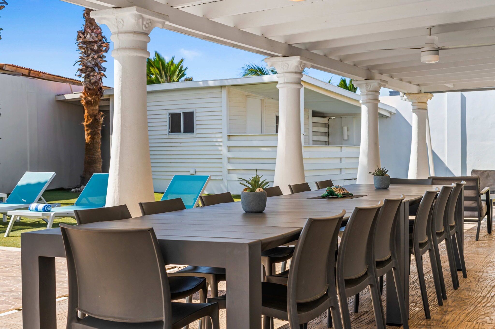 Une terrasse extérieure avec une longue table et des chaises, entourée de colonnes blanches, et un bâtiment blanc en arrière-plan.