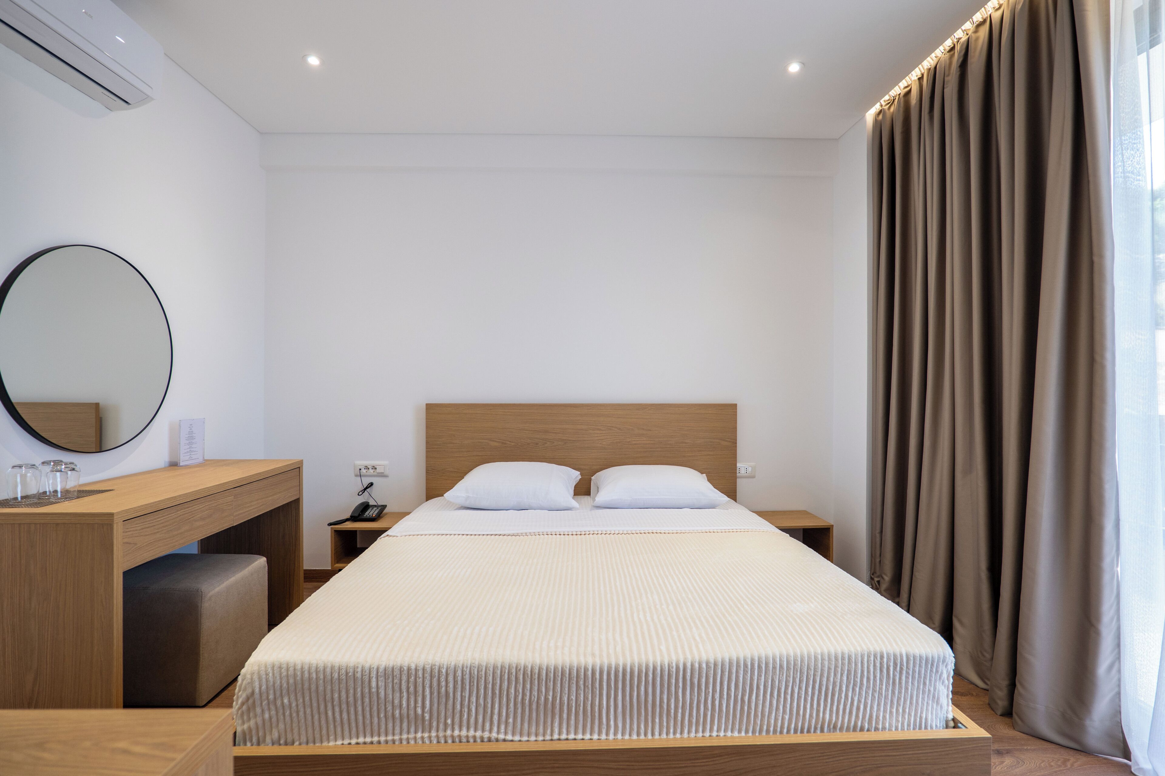 A hotel room with a bed, a desk, a chair, a mirror, a bedside table, and a window with curtains.