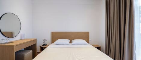 A hotel room with a bed, a desk, a chair, a mirror, a bedside table, and a window with curtains.