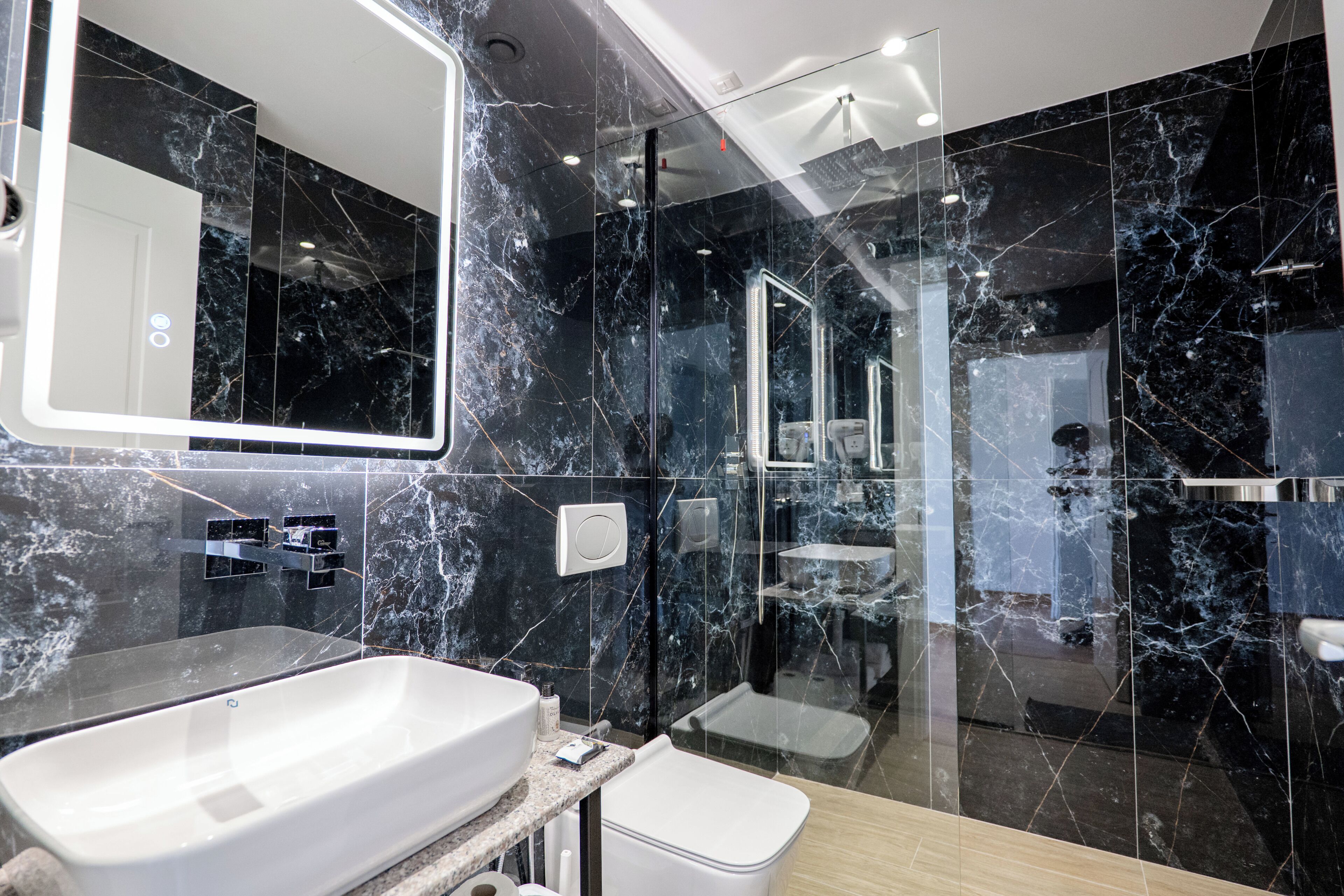 A modern bathroom with a large mirror, a white sink, a glass shower enclosure, and marble walls.