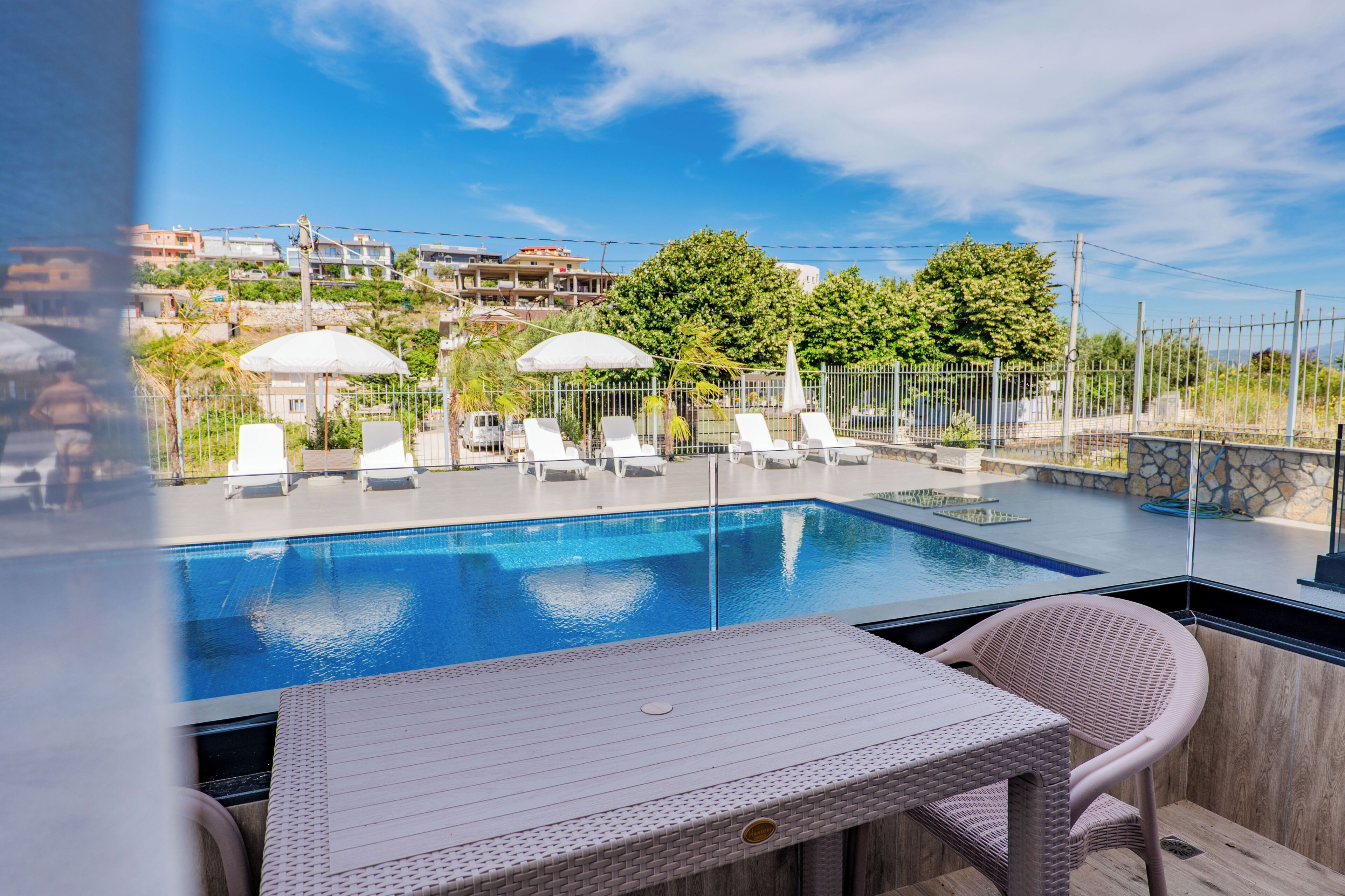 A swimming pool with lounge chairs and umbrellas, a table, and a view of buildings and trees.