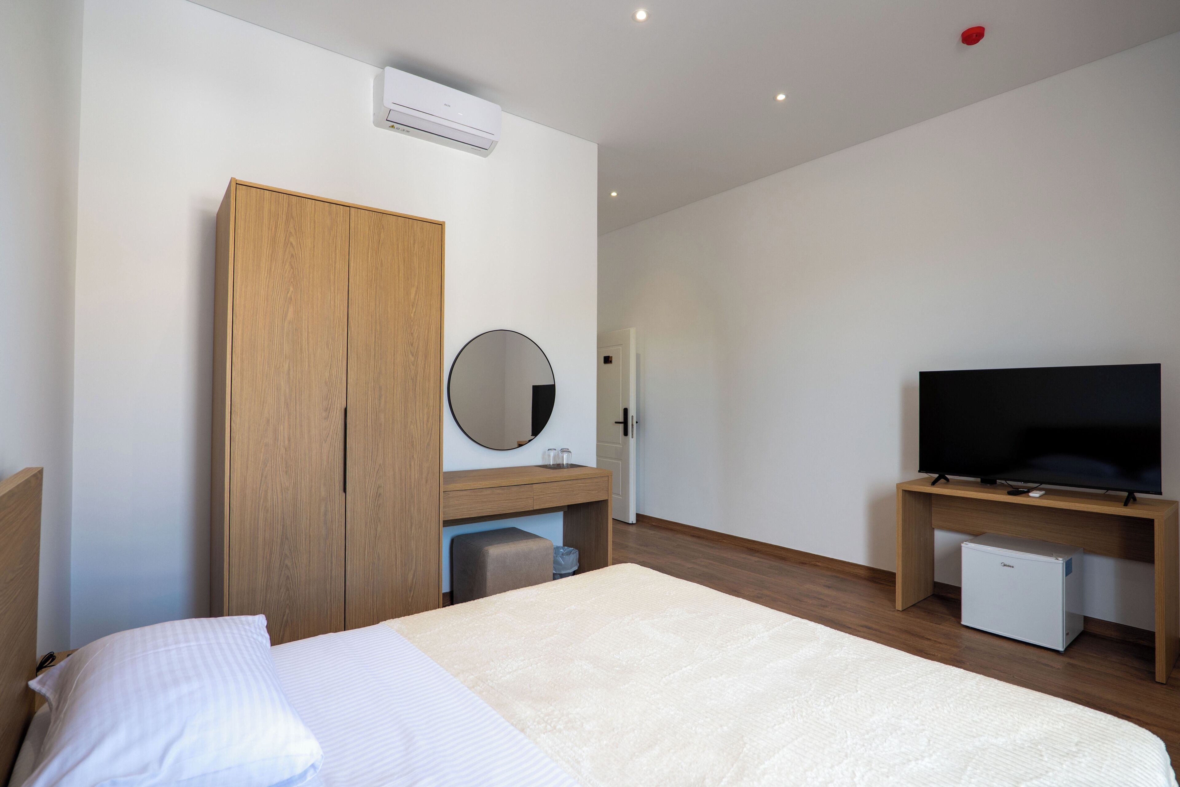 A bedroom with a bed, a wooden wardrobe, a round mirror, a television on a wooden stand, and a small refrigerator.