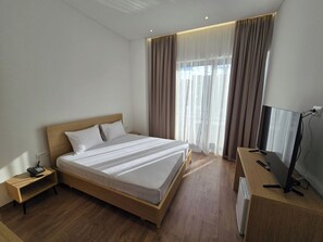 Habitación de hotel con una cama de madera, una mesita de noche con teléfono, un televisor sobre un mueble bajo y un ventanal con cortinas.