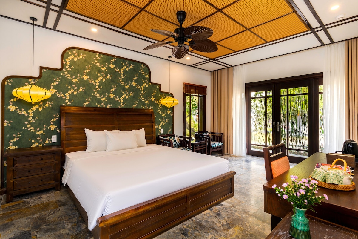 A bedroom with a large bed, wooden headboard, two bedside tables, a desk with a chair, and a window with a view of greenery.
