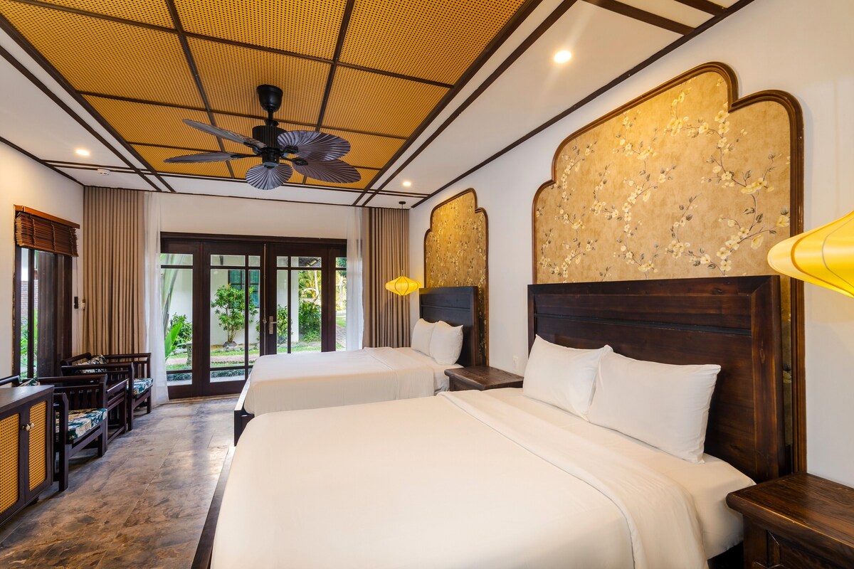 A hotel room with two beds, a ceiling fan, a wooden headboard, a desk with a chair, and a view through the glass door.