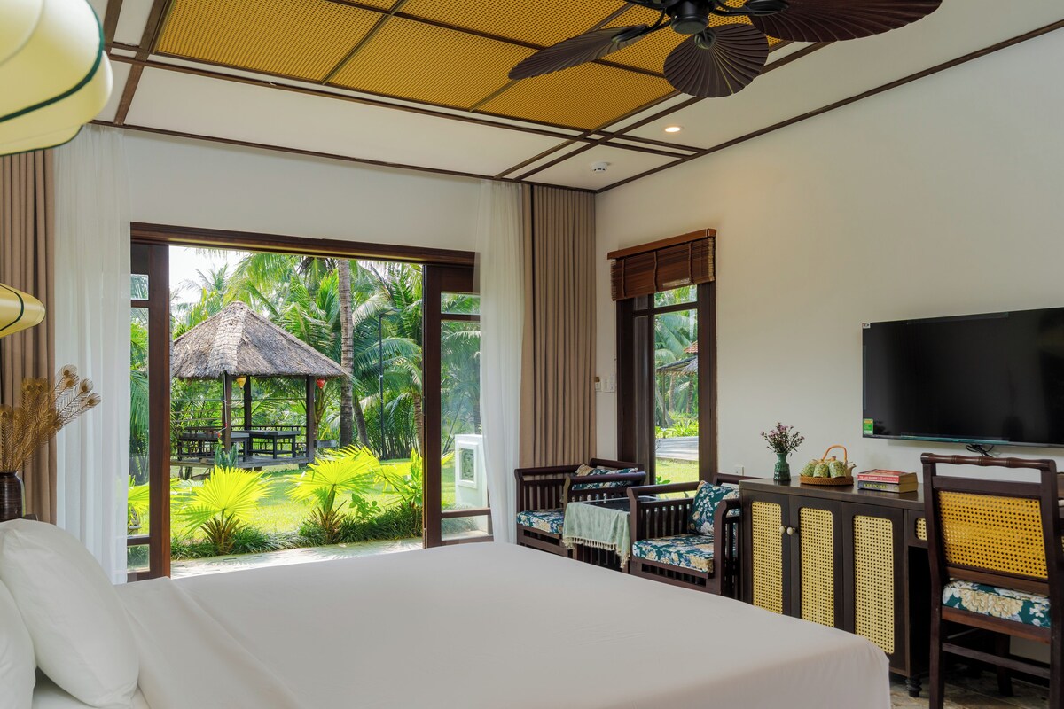 A bedroom with a large bed, a television, and a view of a garden with a thatched-roof structure.