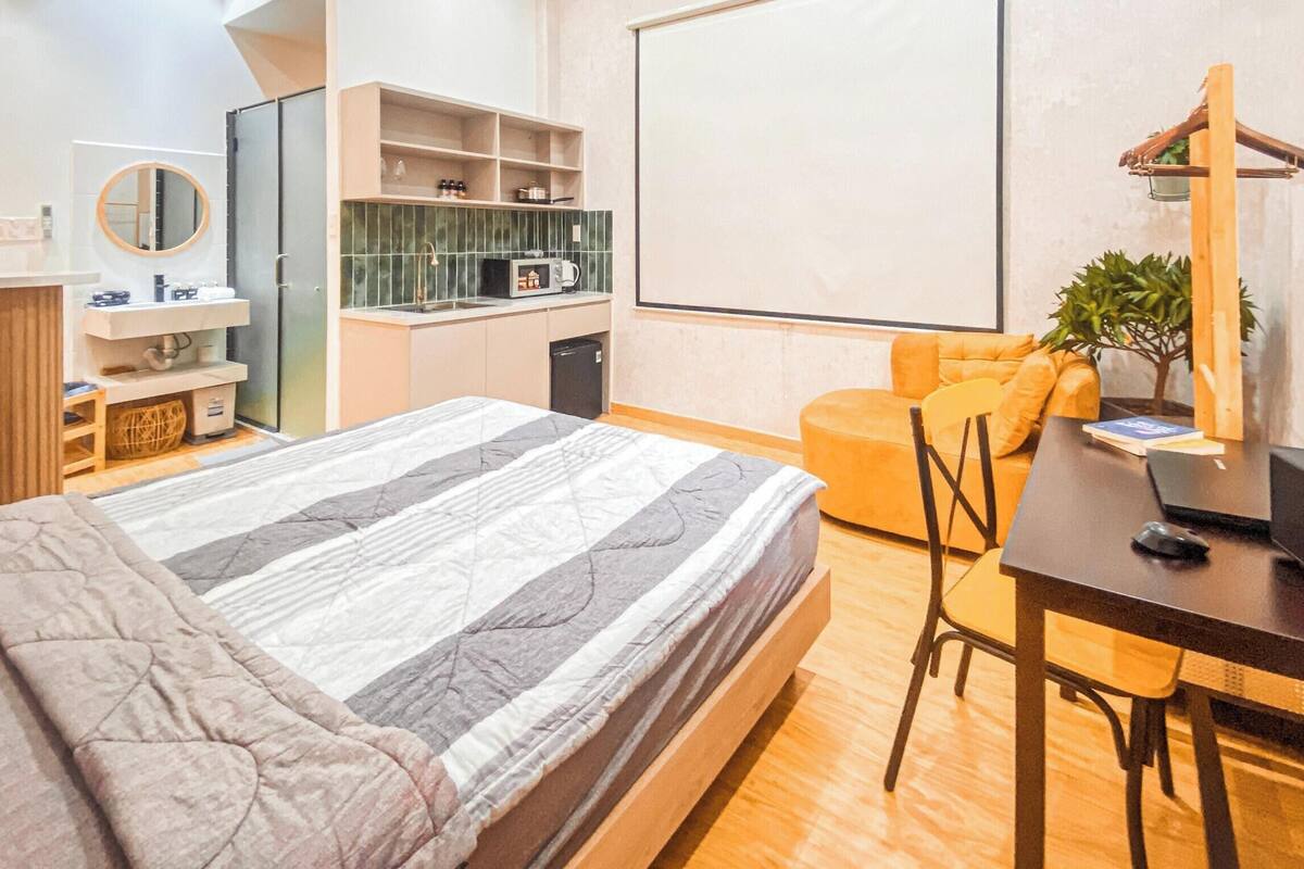 A modern bedroom with a bed, a desk, a chair, a small kitchenette, and a large window.