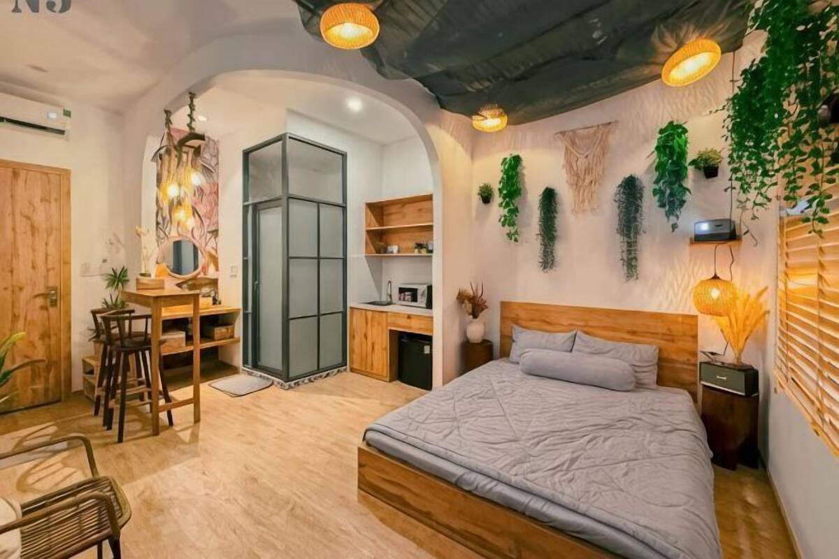 A bedroom with a bed, a desk, and a chair. There is a wardrobe, a mirror, and a shelf with plants.