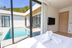 A modern hotel room with a large bed, a TV, and a view of a swimming pool and outdoor seating area. - Aileen Saku Elegant 4BR Villa P3v27/28 (Sa Khu)